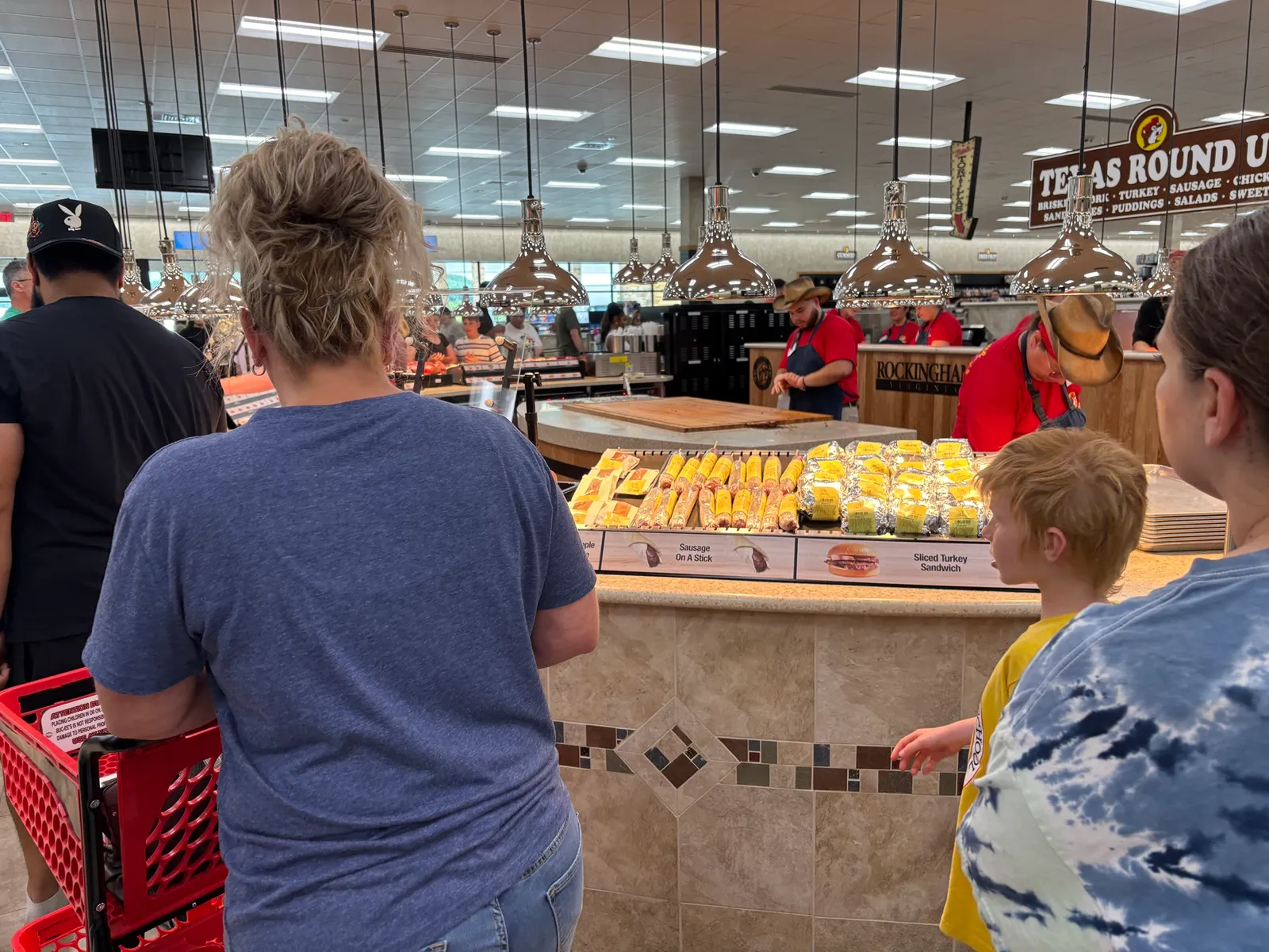 A photo of the interior of a large store. People are waiting in line to get sandwiches which are being kept warm under heat lamps.