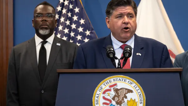 A person stands at a podium with a seal of the State of Illinois, another person standing behind.