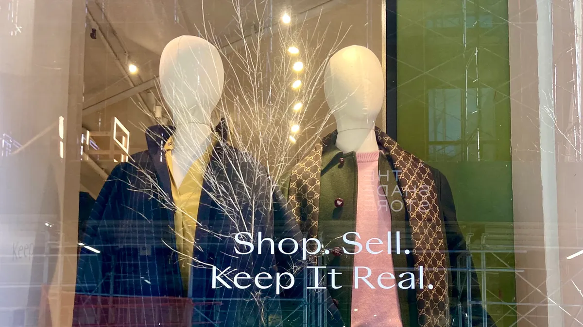 Mannequins flank a wispy decorative branch in a shop window that reads "Shop. Sell. Keep It Real."