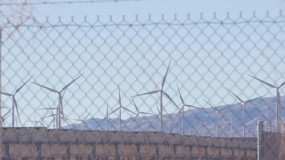 Rows of wind turbines and solar panels viewed through a chain-link fence, with mountains visible in the background under a pale blue sky.