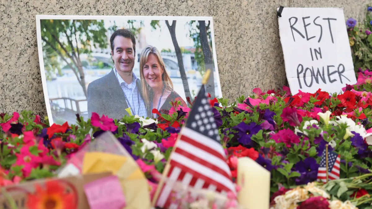 Photo of a couple against a stone wall behind flowers, surrounded by miniature flags and next to cardboard sign that says "REST IN POWER."