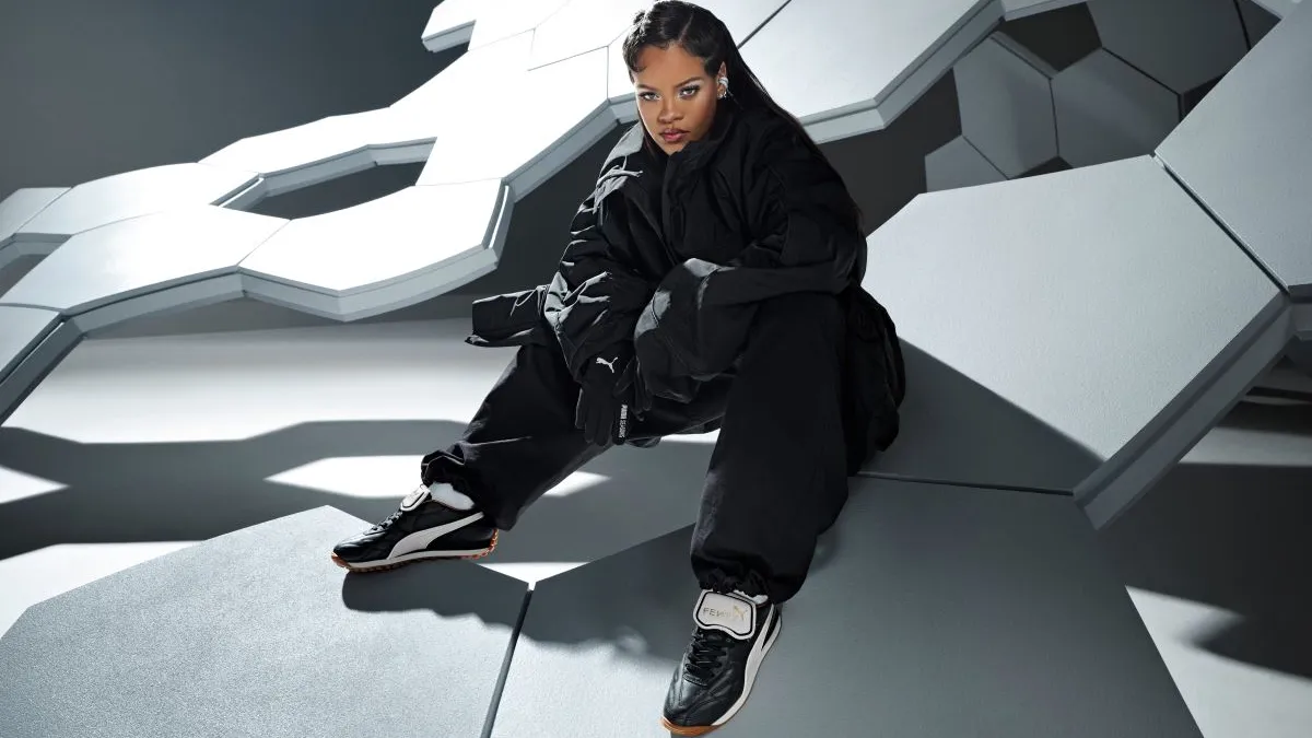 A woman, Rihanna, sits on a giant stylized soccer ball sculpture wearing all oversized black training outfit and Puma soccer shoes