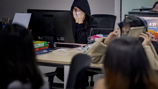 Workers sit at computers on tables in a room, covering their faces with various objects.