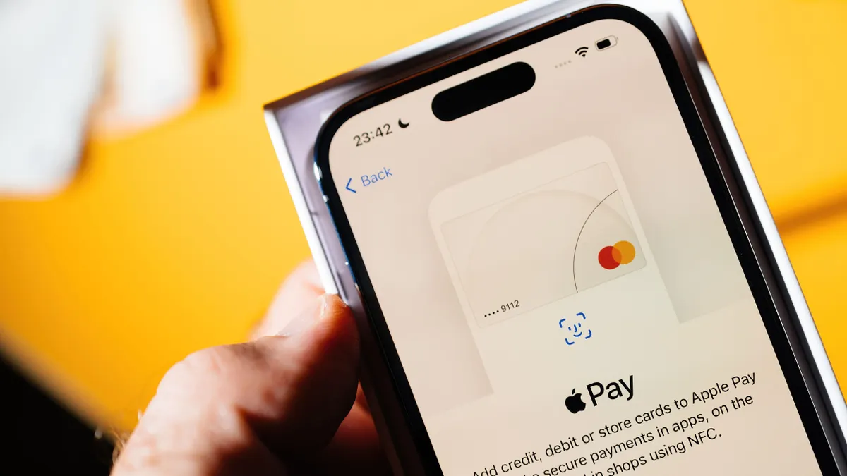 A hand holding an iPhone showing the Apple Pay setup screen. The interface displays a Mastercard graphic with instructions for adding a card, and the phone is partially in its packaging against a warm yellow background.