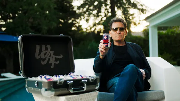 Charlie Sheen sits outside holding an aluminum can next to a suit case full of the cans.