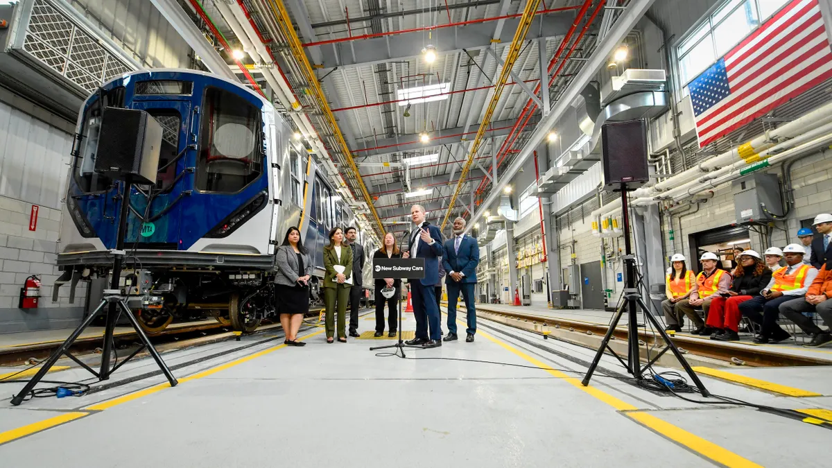 Several people stand at a podium in a large, brightly lit industrial building next to  a subway car on rails.