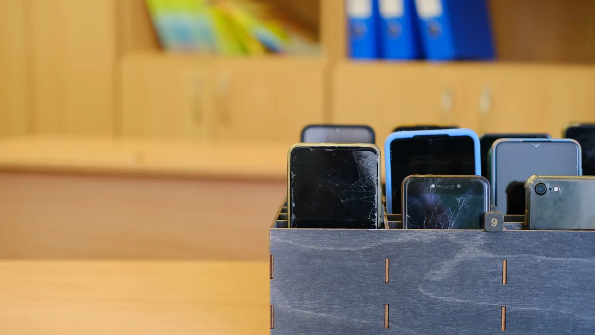 A hanful of cellphones sit in a wooden box in rows on top of a desk in a room.