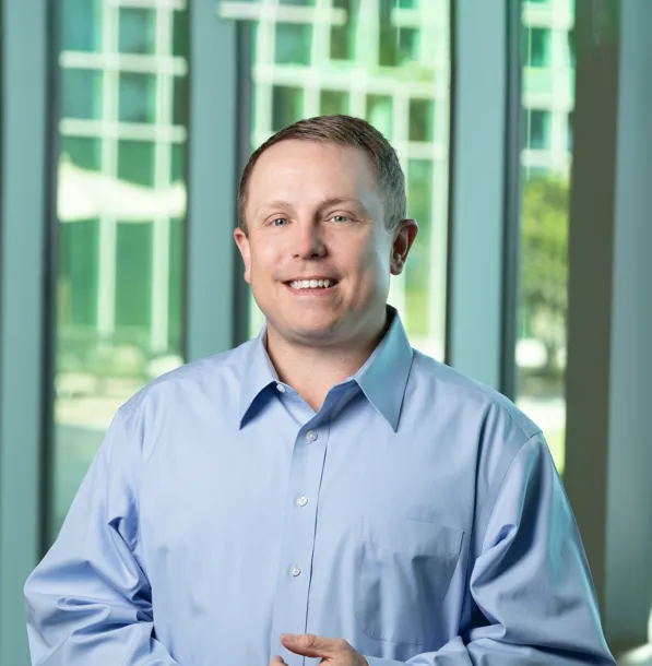 Headshot of a white man in a light blue button up shirt standing in front of a large glass window