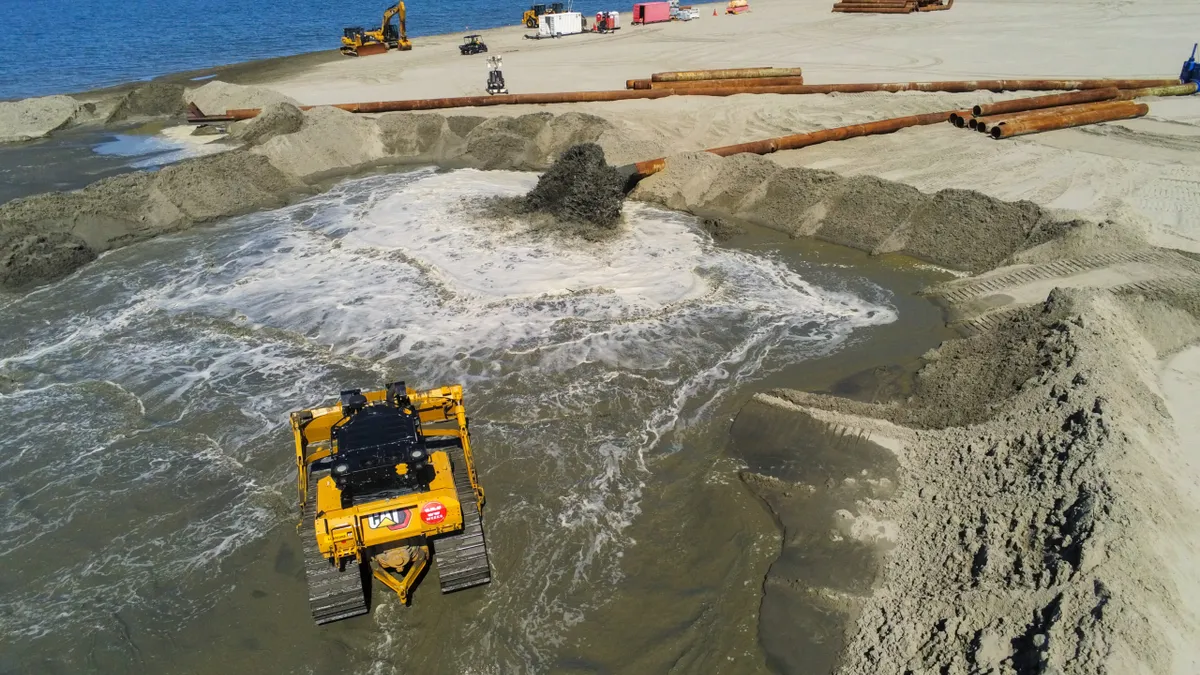 Yellow excavation equipment digs a wide trench on a beach, with massive pipes stacked in the background.