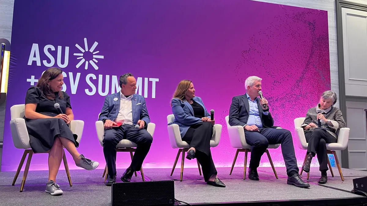 Five people sit in chairs on a stage, each with a microphone, taking turns speaking on a panel.