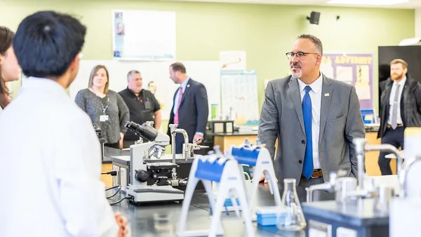 A person is standing at a side angle in a classroom with laboratory tables facing another person whose face is turned to the other person. Other people are standing around in the background