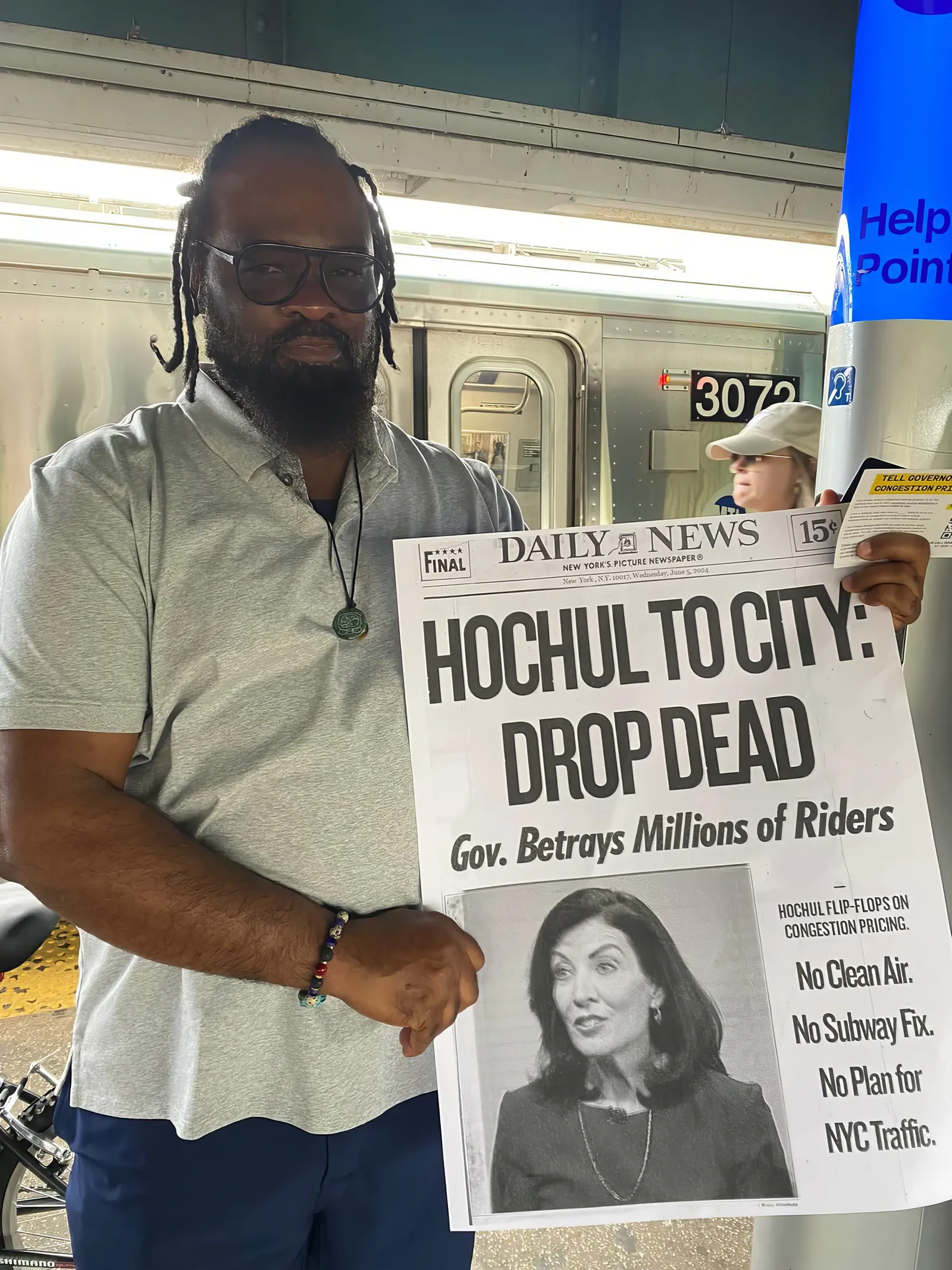 A man in a New York subway car holding a poster of a newspaper front page that reads, &quot;Hochul to city: Drop dead.&quot;