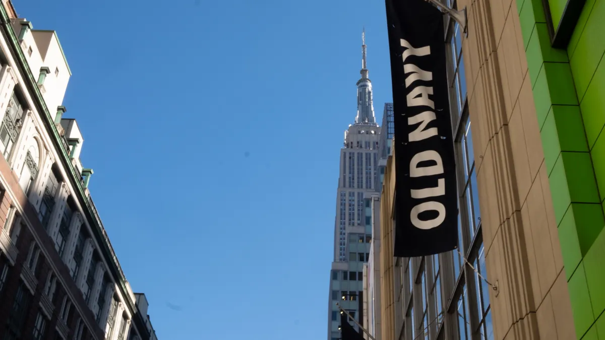 A dark blue flag that reads "Old Navy" in white block letters hands vertically on a building in New York City, shown in the righ of a photo. Another building is on the left, with blue sky in the middl