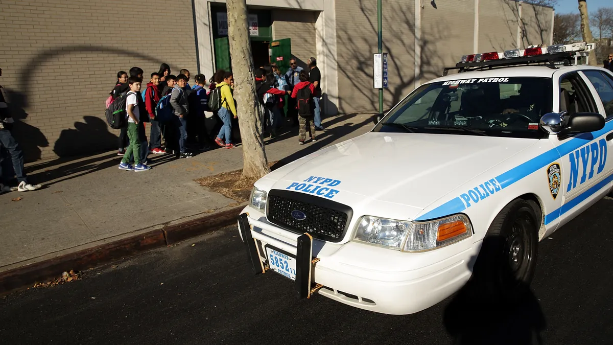 An NYPD police car is parked outside of a school with students lined up in the background