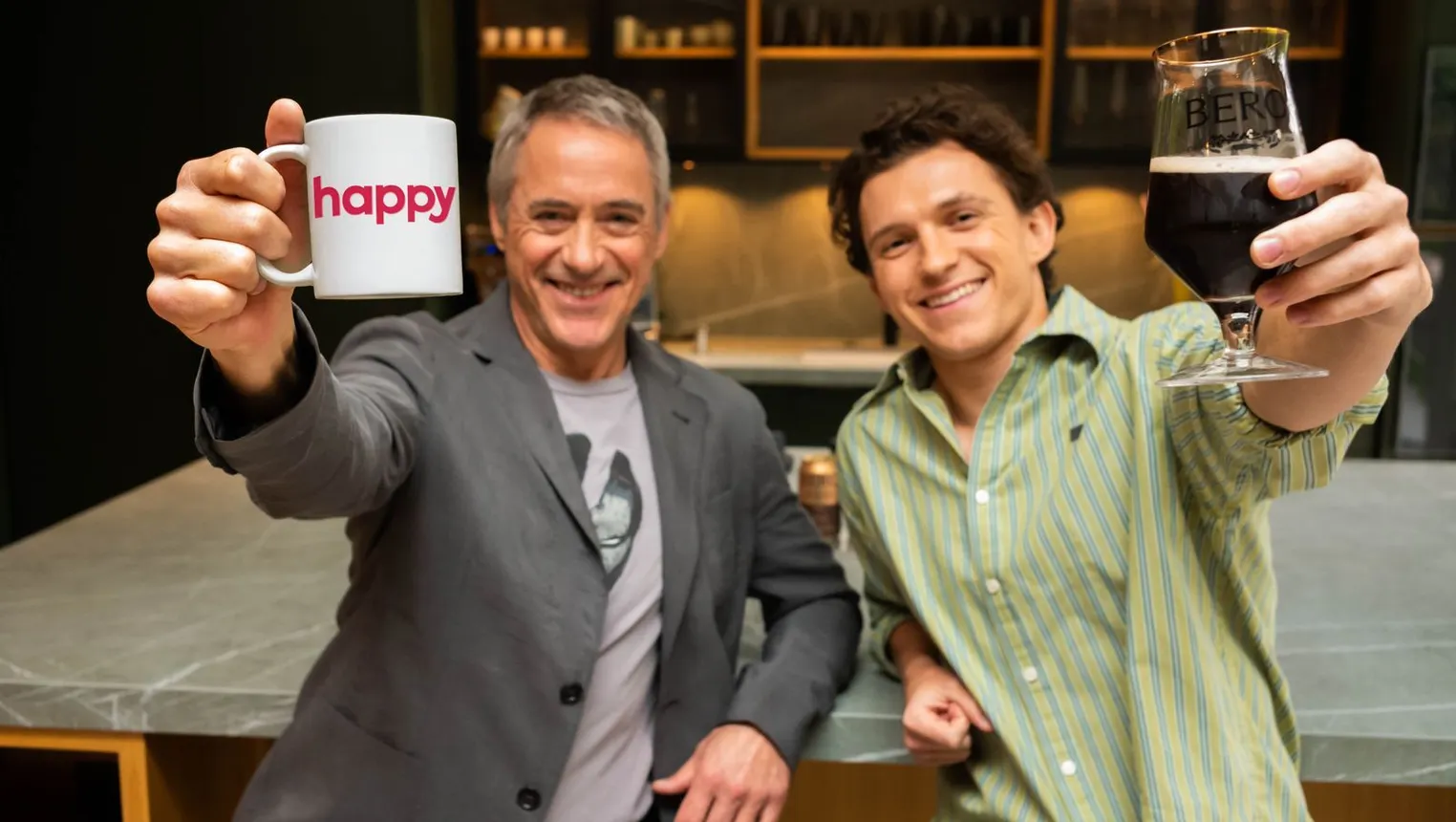 Actors Tom Holland and Robert Downey Jr. sit next to each other raising a glass of beer and coffee, respectively.