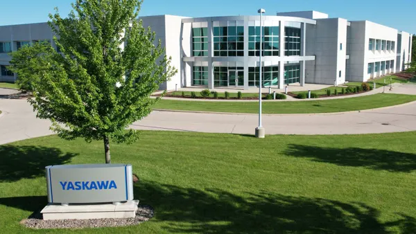 A blue and silver Yasakawa sign in front of a tree, with a white building with windows in the background.
