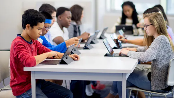 A group of high school students sit together at a long table while individually using digital tablets.