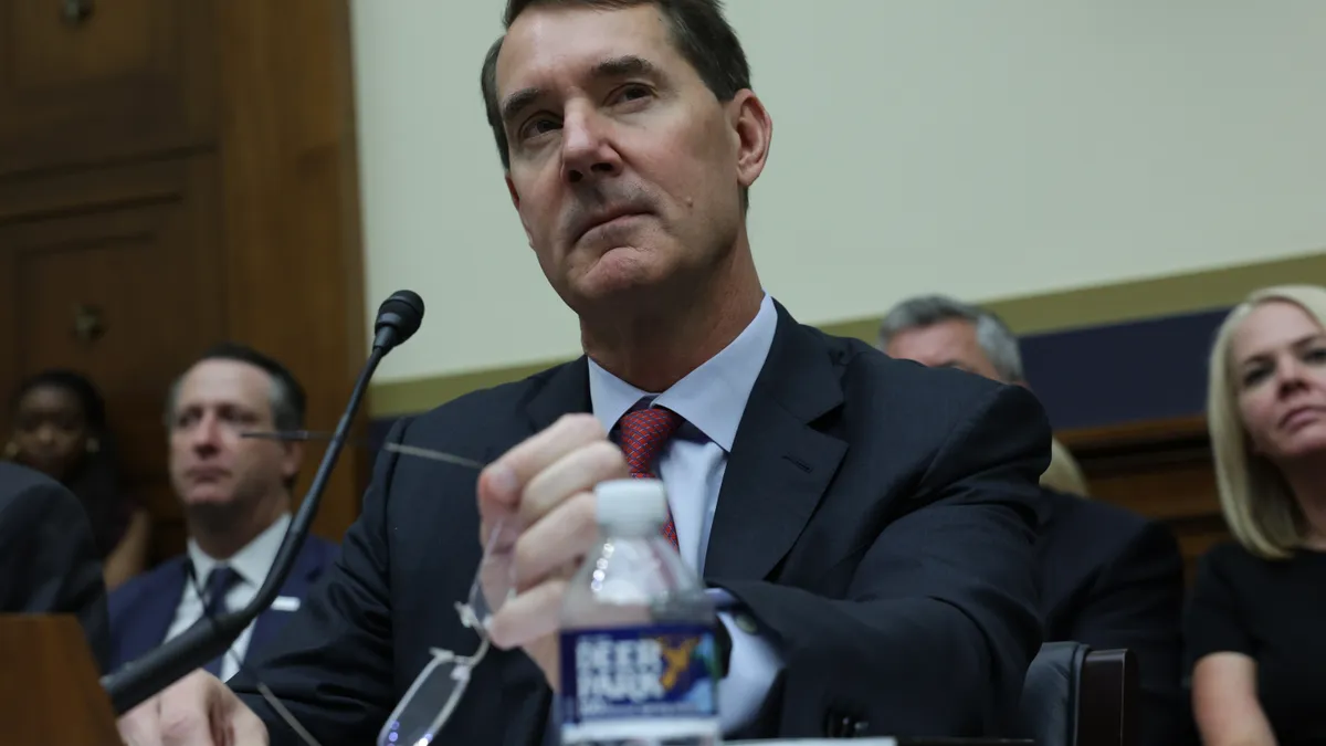 PNC CEO Bill Demchak holds his glasses and looks on during a Congressional hearing, as people sit behind him in the background.