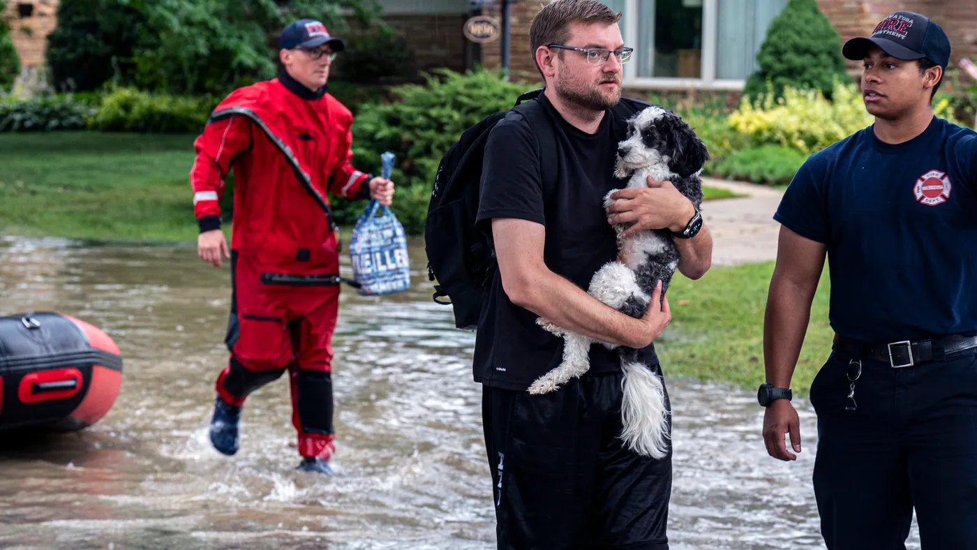 A person carrying a wet dog wades through a flooded street while two people look on.