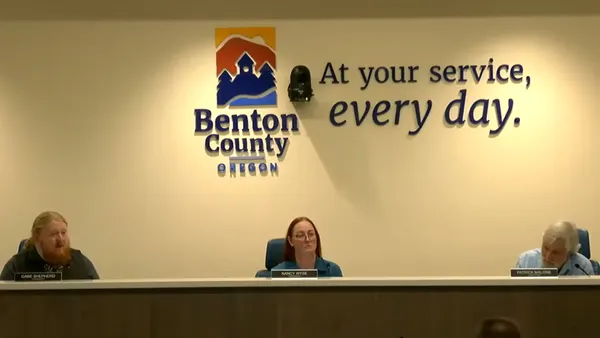 Three people sit at a long dais with the Benton County, Oregon, seal and the slogan "At your service, every day," behind them.