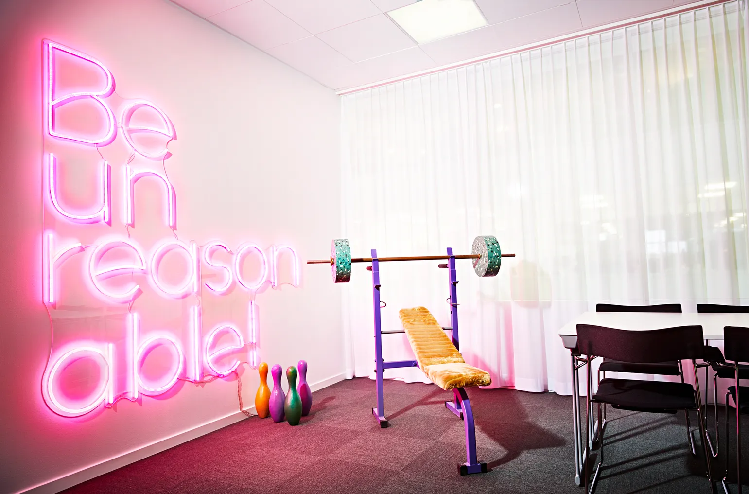 Room with a neon sign that says 'Be Unreasonable' with weights and a desk.