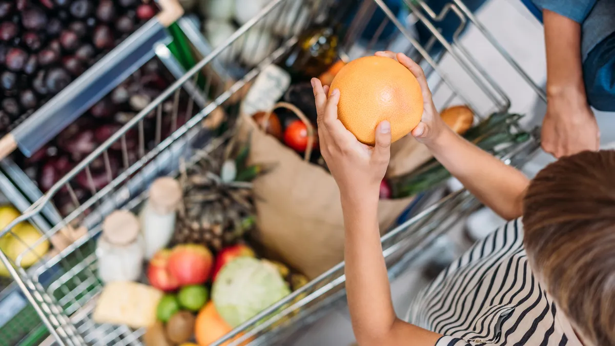 Overhead view of a child helping their parent grocery shop by placing a piece of fruit into a shopping cart.