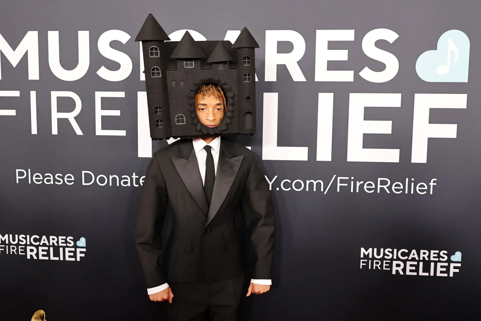 A person in a black suit wears a castle shaped headdress and stands on a red carpet.