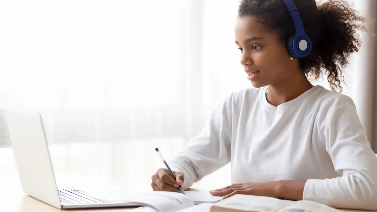 African American teen girl wearing headphones learning language online, using laptop, looking at screen, doing school tasks at home, writing notes, listening to lecture or music, distance education