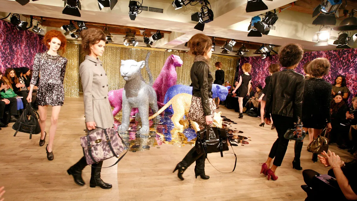 Models with bouffant hairdos walk a runway holding large handbags.