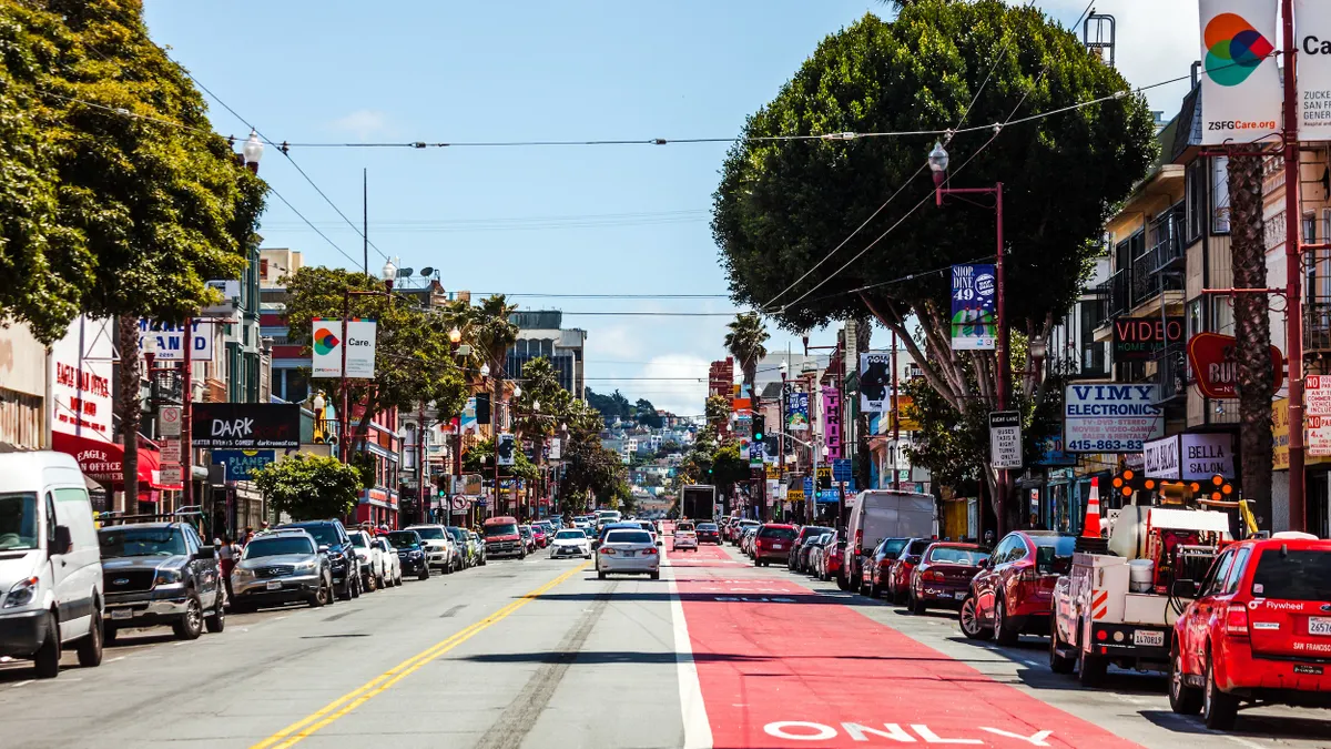 View down a two-lane street with a red-painted bike line on the right and parked cars on both side of the street, which is lined with small shops.
