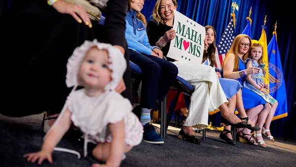 A woman holds a sign that says "MAHA Moms" as she sits on stage