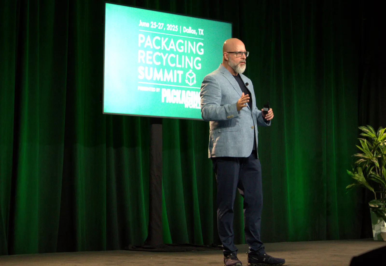 A person stands on a stage in front of a green background that says Packaging Recycling Summit and speaks to an audience.