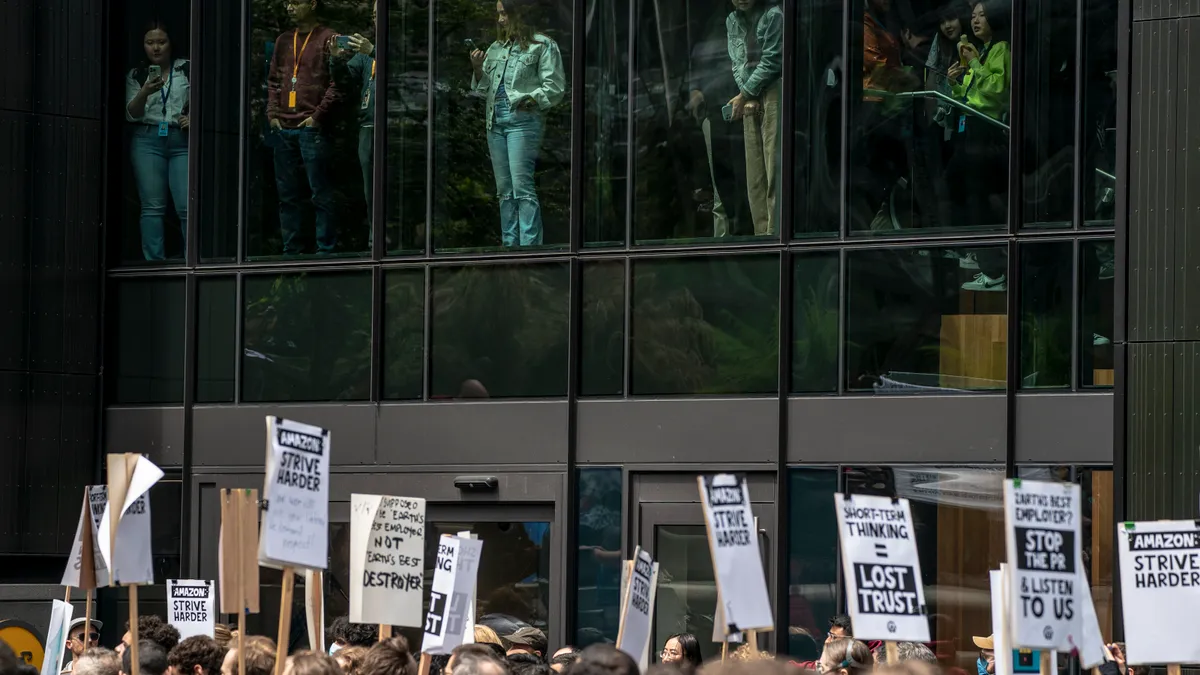 The heads of a group of protestors can be seen along with signs, walking past a building with workers looking out from inside.