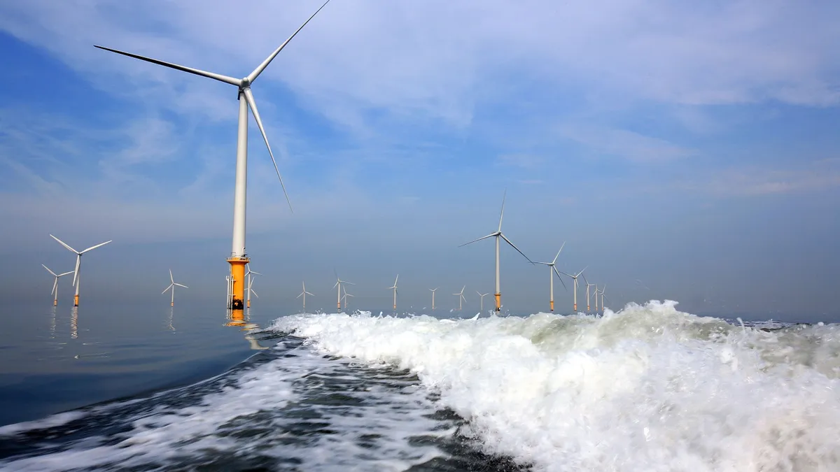 About a dozen white wind turbines stand in the ocean as waves crash around their bases.