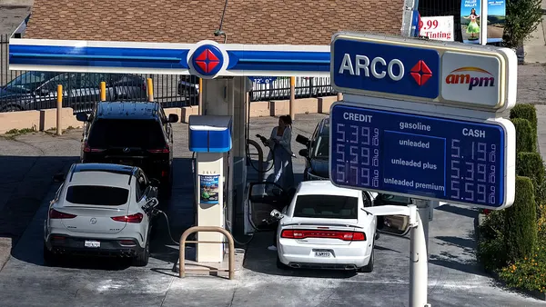 Two cars fill up at a gas station, with prices posted over $5 a gallon.