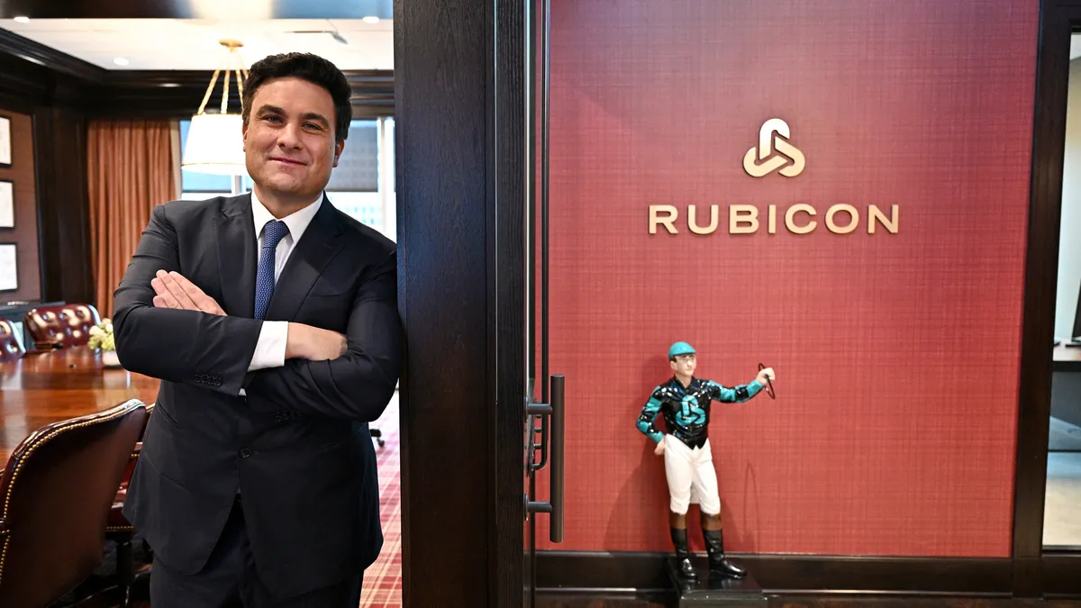 Man in suit standing in office, with statue of horse jockey and Rubicon logo to the side