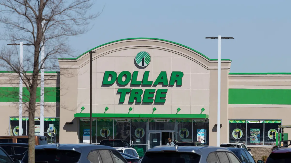 A Dollar Tree store with a large green and white sign above the entrance, located in a shopping center. The building has a beige exterior with green trim and large windows displaying promotional signs. Several parked cars are visible in the foreground, and a leafless tree stands on the left side of the image under a clear blue sky.