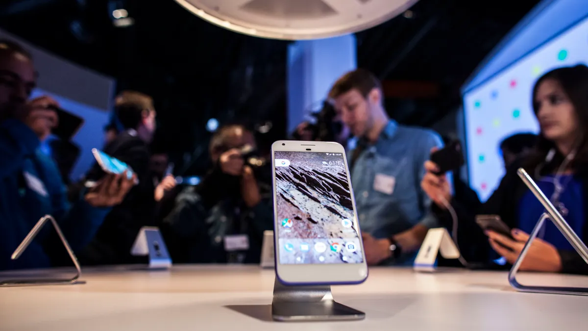 A Google Pixel phone on a table in the foreground with people in the background examining Google devices.