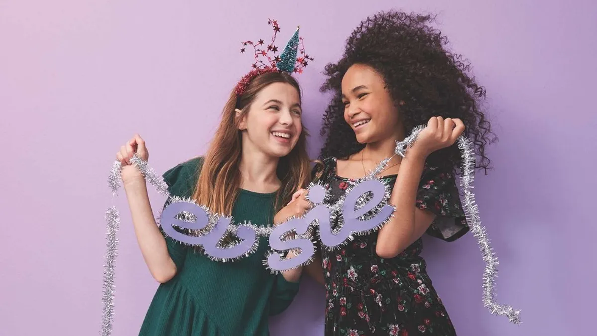 Two tween girls stand next to each other in dressy clothes. One wears a party hat, and they hold an Evsie banner.