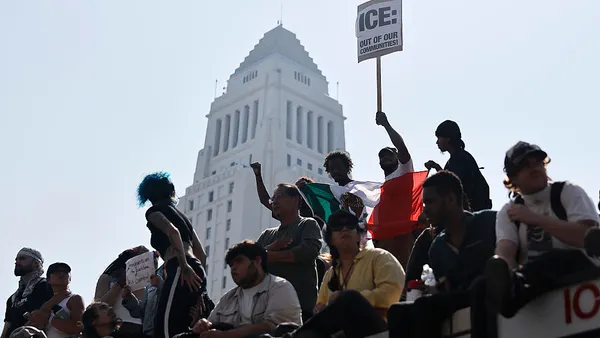 A group of people sit on a bridge in front of a tall white building, one holding a sign that says "ICE out of our communities."