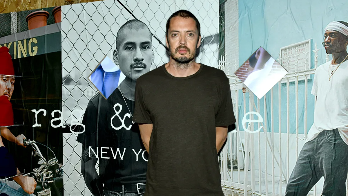 A man in a dark t-shirt, jeans, and short hair stands unsmiling in front of a photo collage backdrop featuring images of models wearing casual clothing.