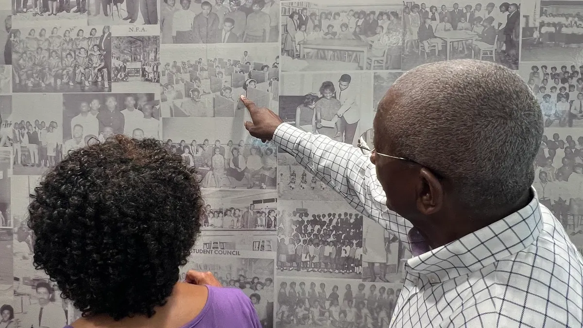 A person stands next to someone while pointing to one of the many black-and-white photos displayed on a wall.