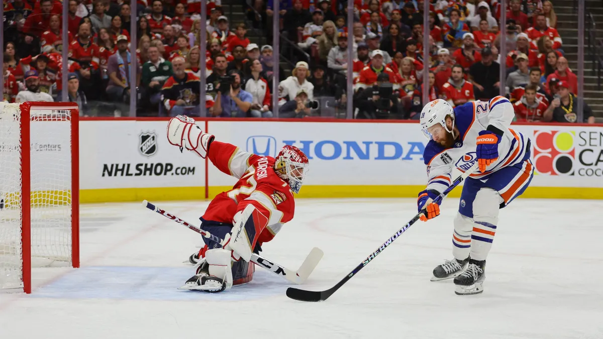 A hockey player scores a goal while another attempts to block while a crowd watches in the background.