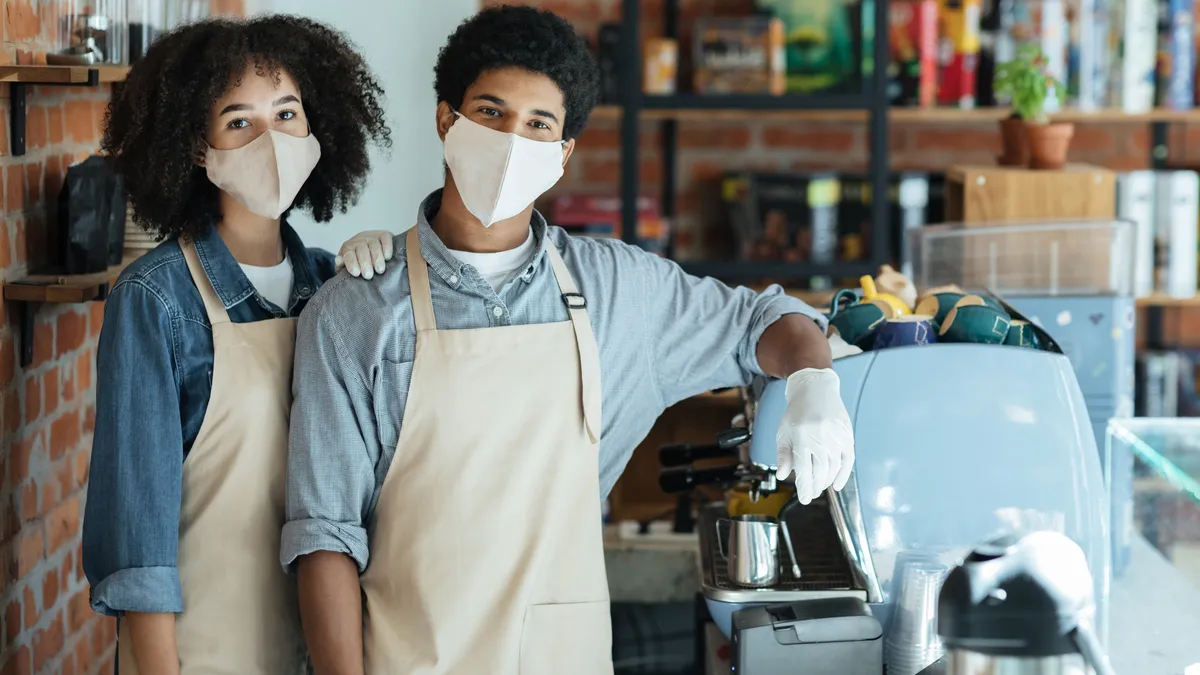 Family small business and work during COVID-19 epidemic. Millennial african american couple in apron, protective mask and gloves look at camera in interior of modern coffee shop with equipment