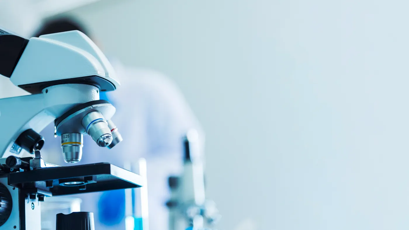 A laboratory microscope sits beside test tubes as a researcher works in the background.