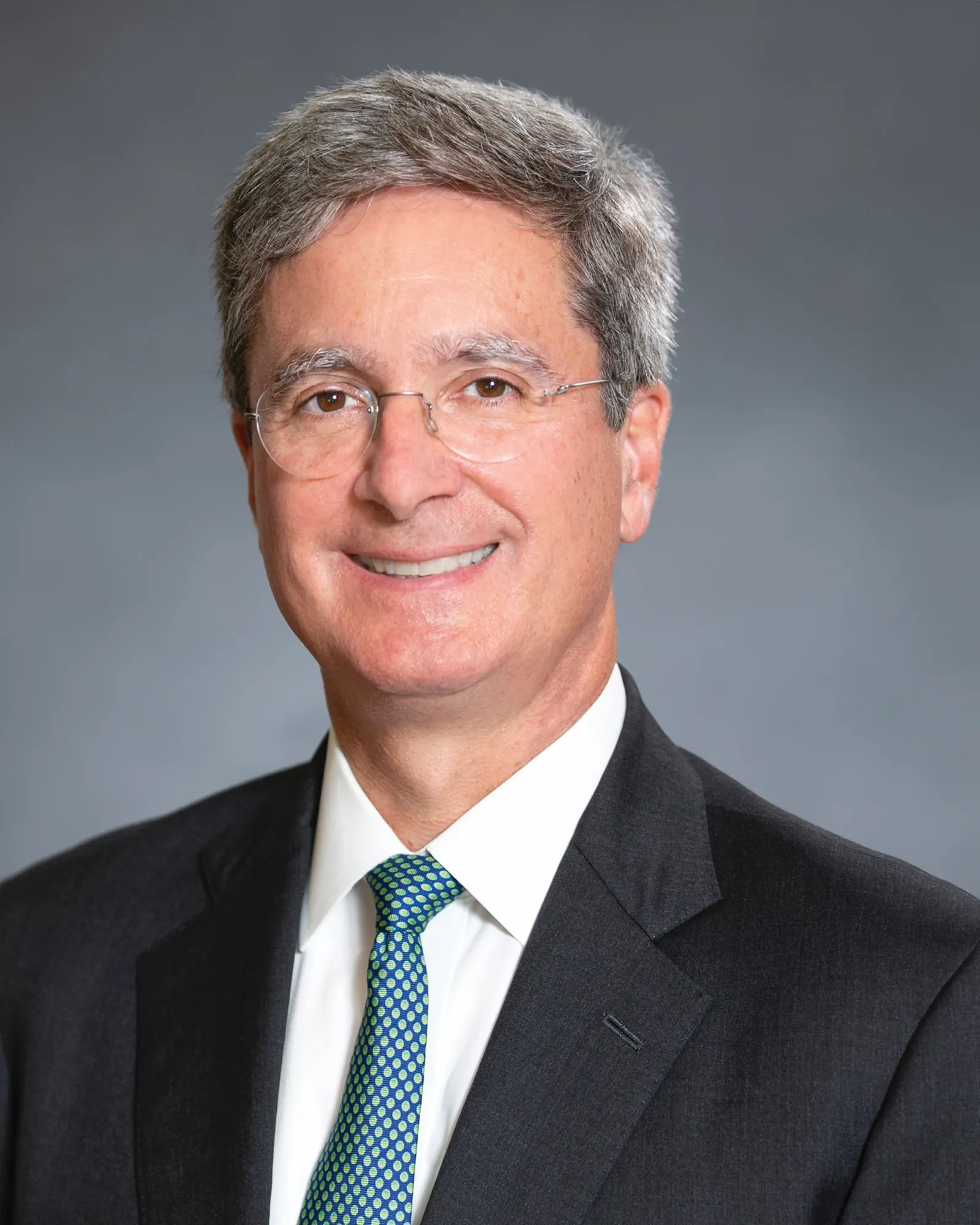 A person wearing glasses and a suit and tie smiles for the camera.
