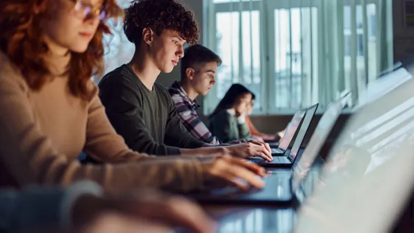 A group of students works on their laptops while seated at desks in a classroom.