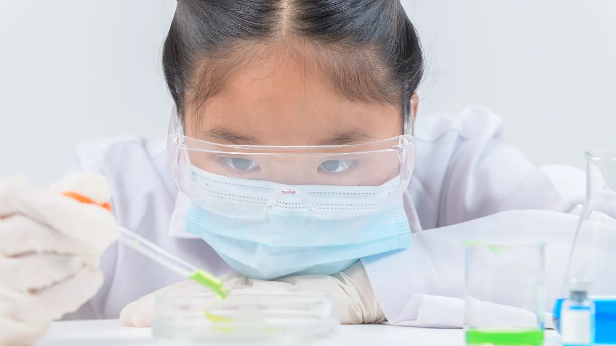 Girl wearing safety glasses and lab coat making experiment and using dropper in chemical laboratory, education and science concept