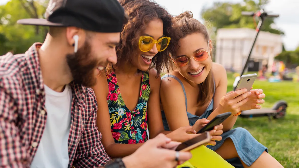 Three young adults sit on grass in a sunny park, smiling and looking at their smartphones