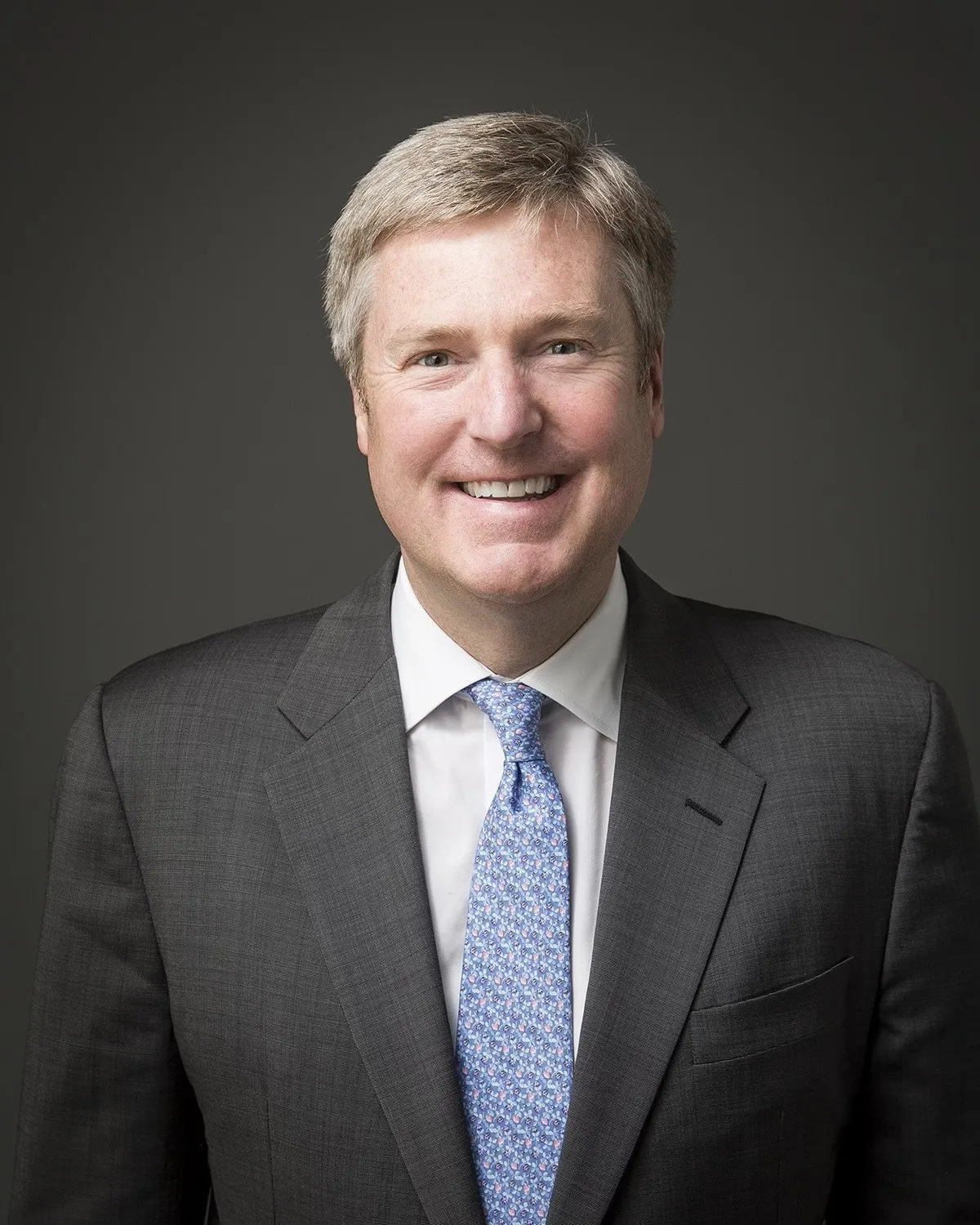 Headshot of advertising executive John Osborn wearing a charcoal suit, blue patterned tie and white shirt against a dark gray background.